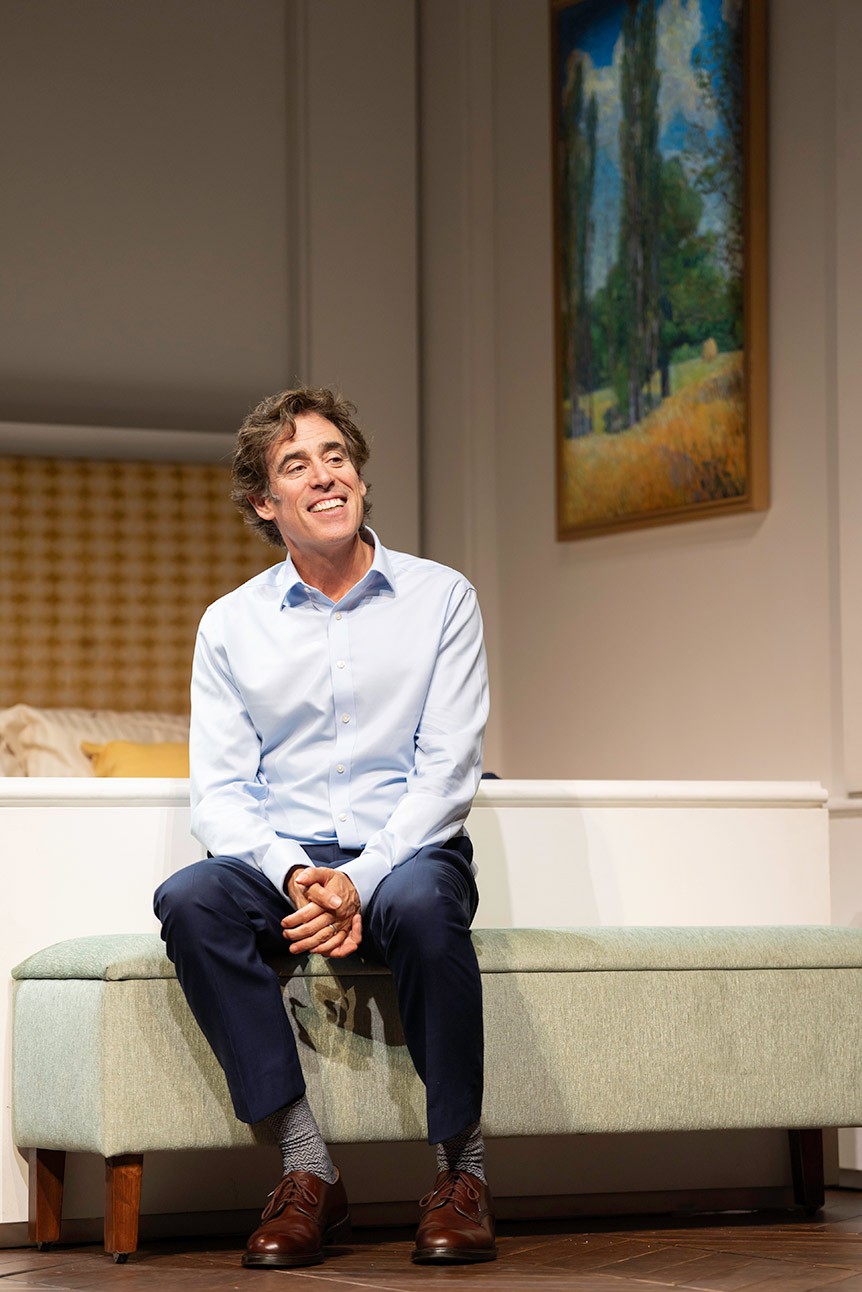 A man in a light blue shirt sits smiling on a light green sofa; painting in background.