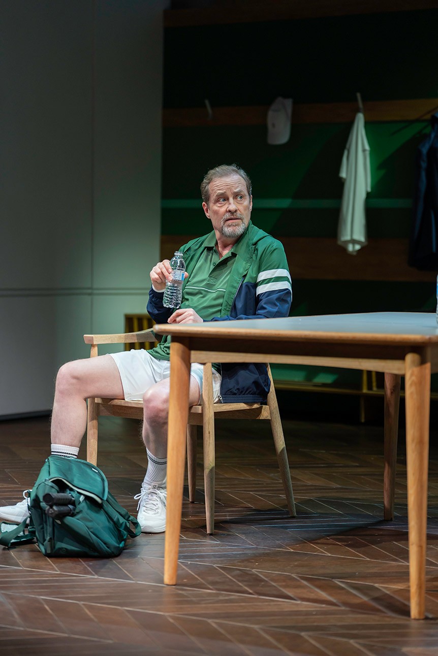 Man in green sports attire sits at table, holding a bottle, with a gym bag nearby.