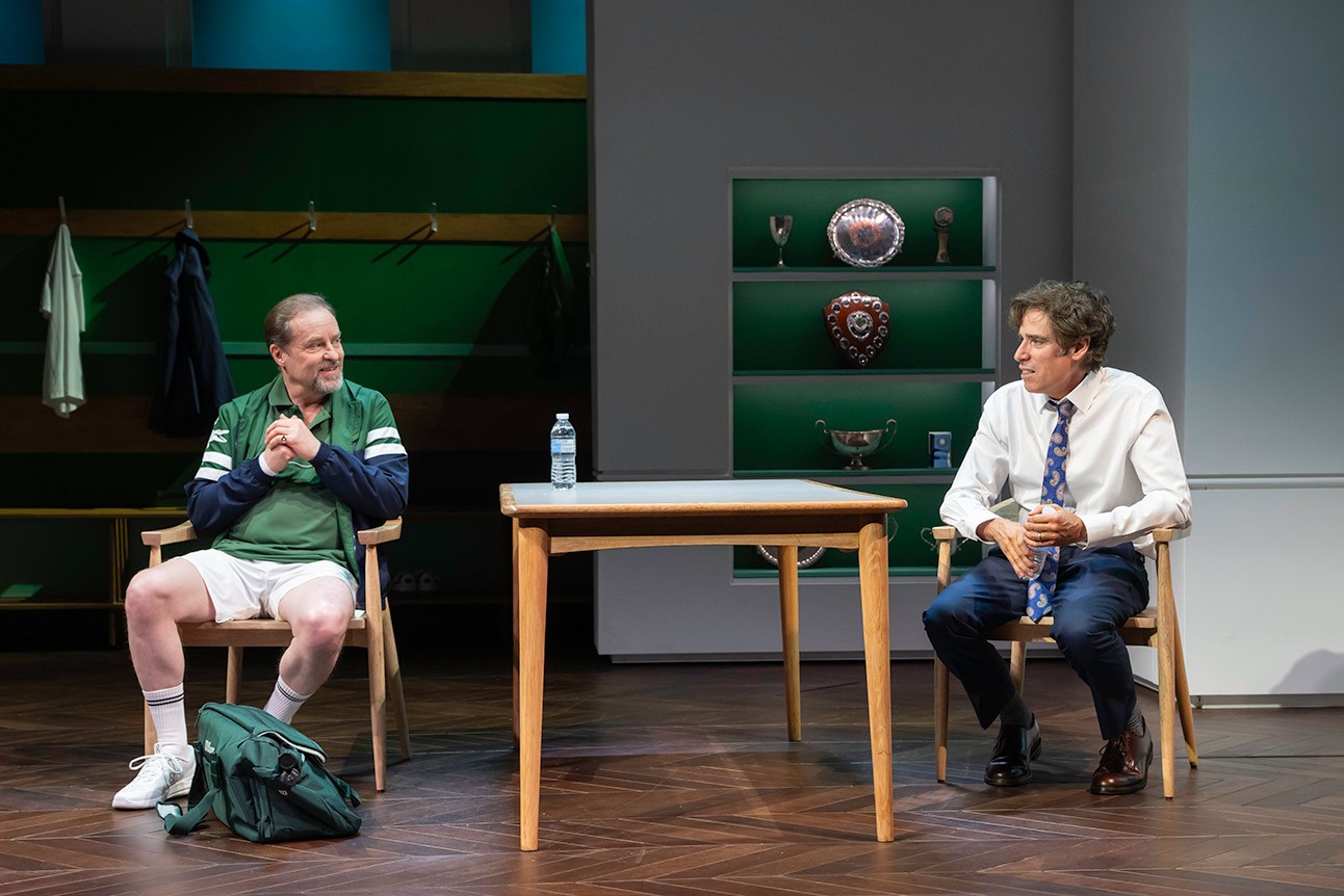 Two men seated on stage, one in green sports attire, the other in a suit, conversing.