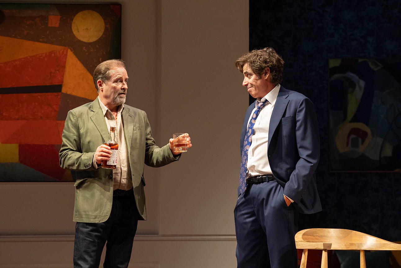 Two men in suits conversing on stage; one holds a drink, abstract art in background.