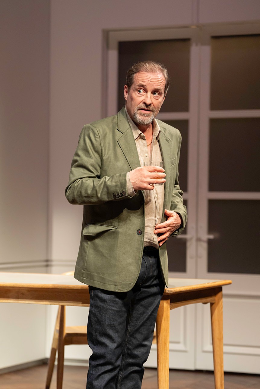 Man in green jacket gestures while speaking, standing near a wooden table in a bright room.