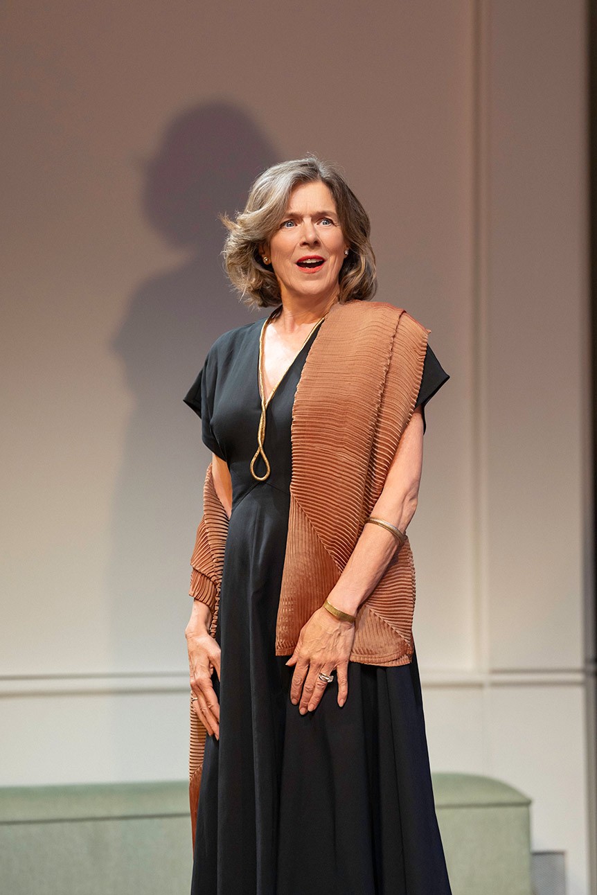 Woman in black dress and brown shawl, speaking on stage, with neutral background.