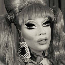 A person with dramatic makeup, large earrings, and styled hair, posing against a neutral background.