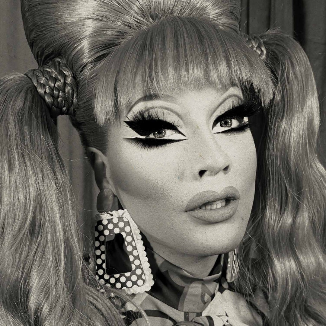 A person with dramatic makeup, large earrings, and styled hair, posing against a neutral background.