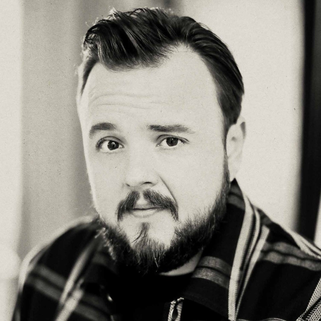 A man with a beard and short hair, wearing a checked shirt, looks directly at camera.