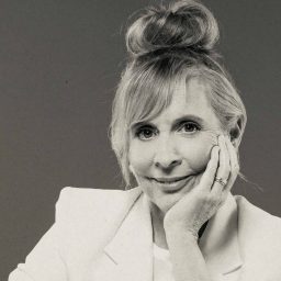 A woman with a top bun hairstyle, wearing a light blazer, smiles with hand on cheek.
