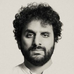 A person with curly hair and a beard looks directly at the camera against a plain background.