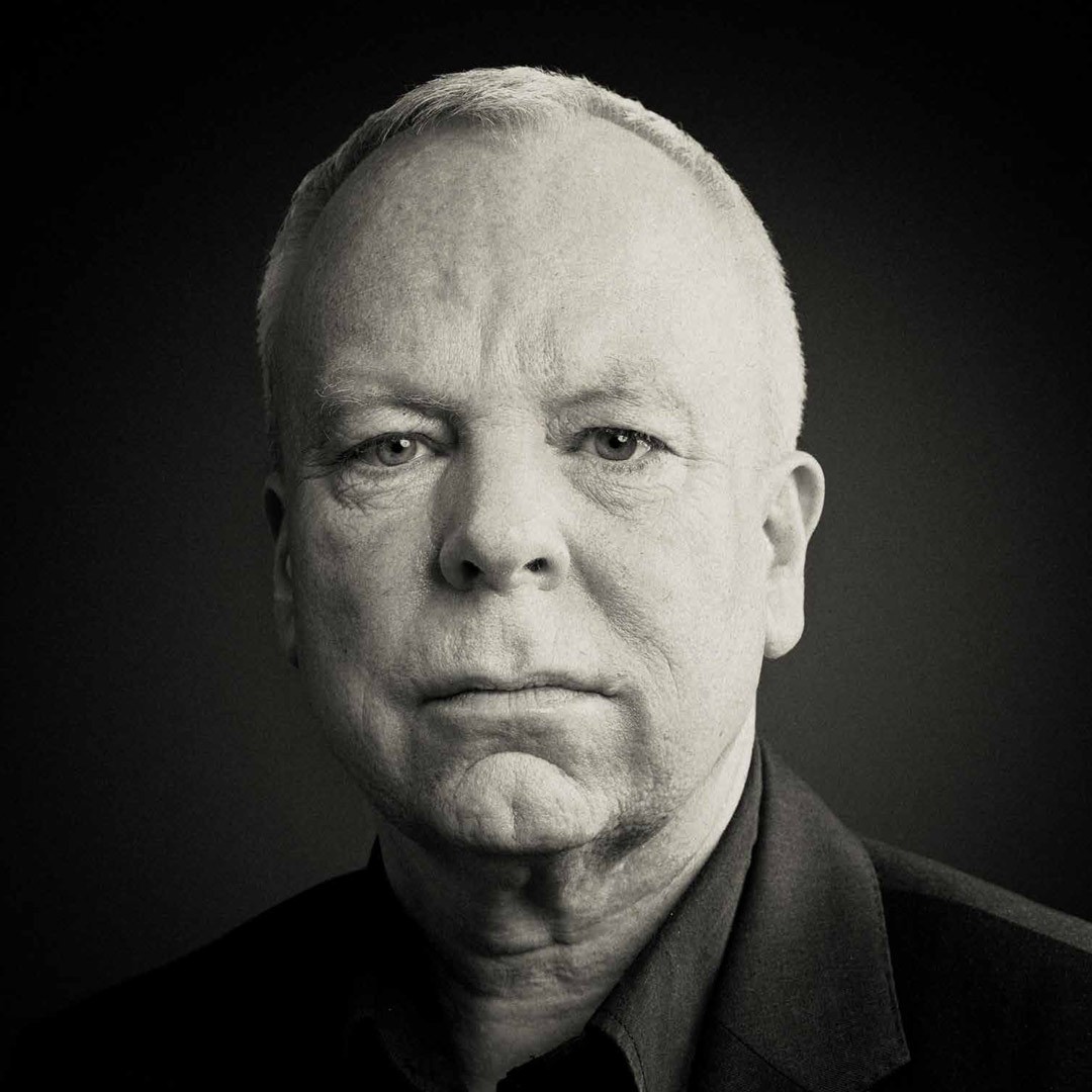 A man with short hair and a serious expression, wearing a dark shirt, monochrome background.