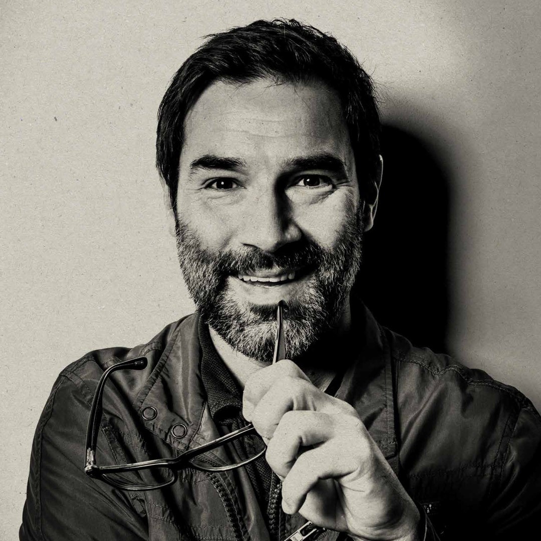 Man smiling, holding glasses near his mouth, monochrome image with textured background.