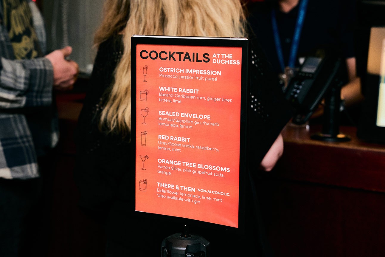 A person with long blonde hair stands near a red cocktail menu at an event.