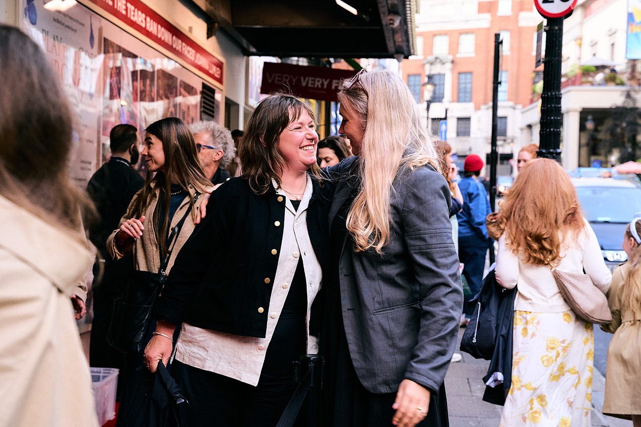Two women smiling and conversing outside a theatre, surrounded by a crowd, urban background.