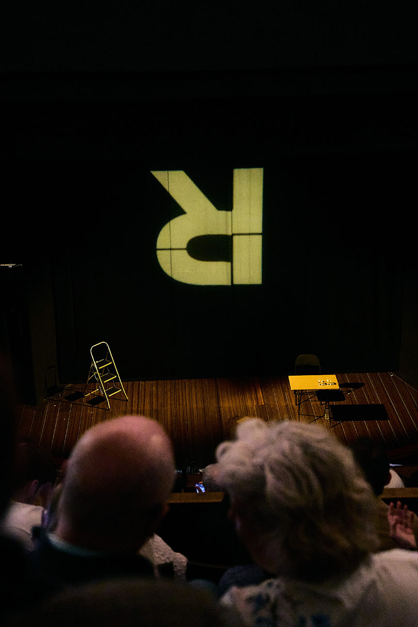 Stage with ladder, table, and large inverted "R" projection; audience heads visible in foreground.