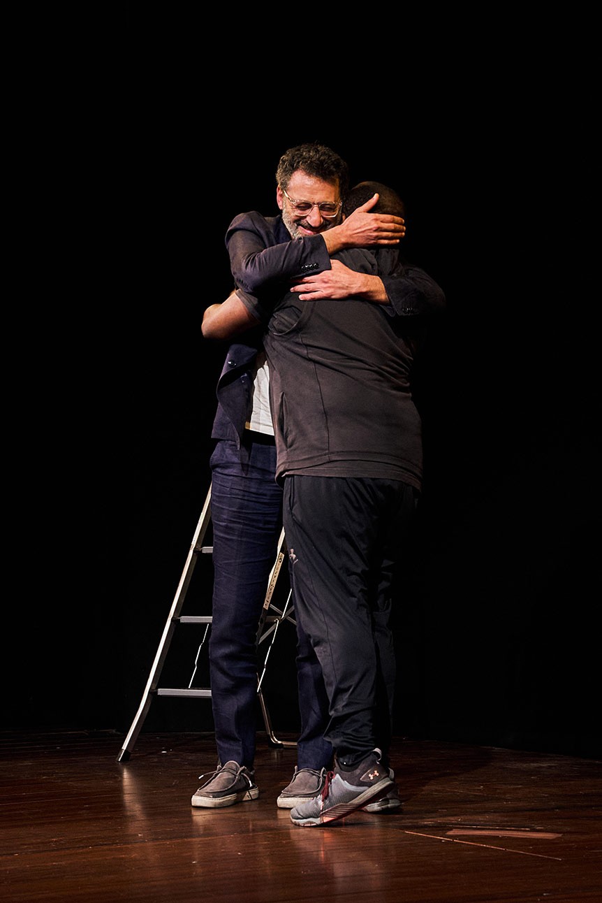 Omar Elerian & Lucian Msamati embrace on the Duchess stage, with a ladder in the background against a dark backdrop.