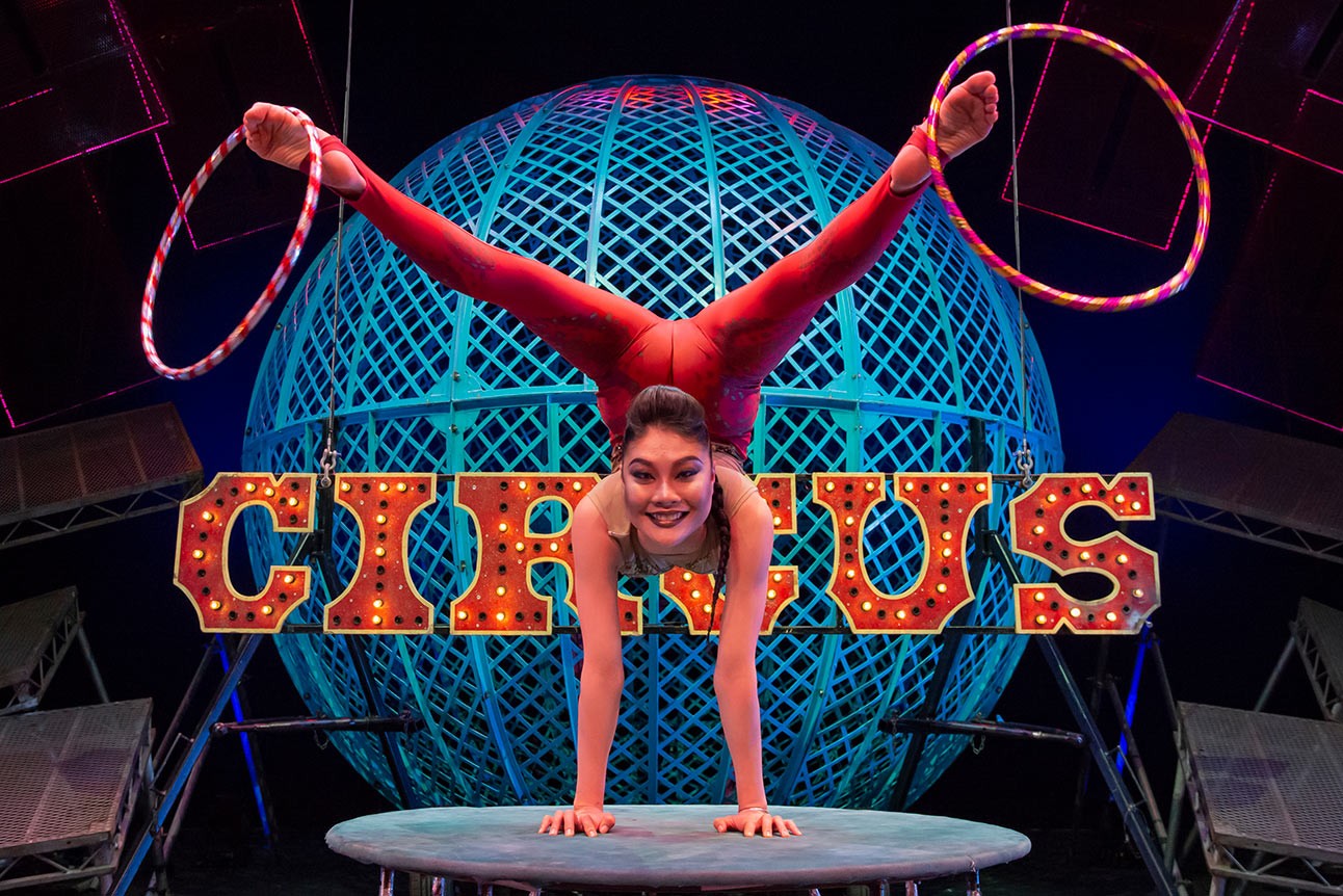 Performer Elberel in red costume doing handstand splits on stage, with "CIRCUS" sign behind.