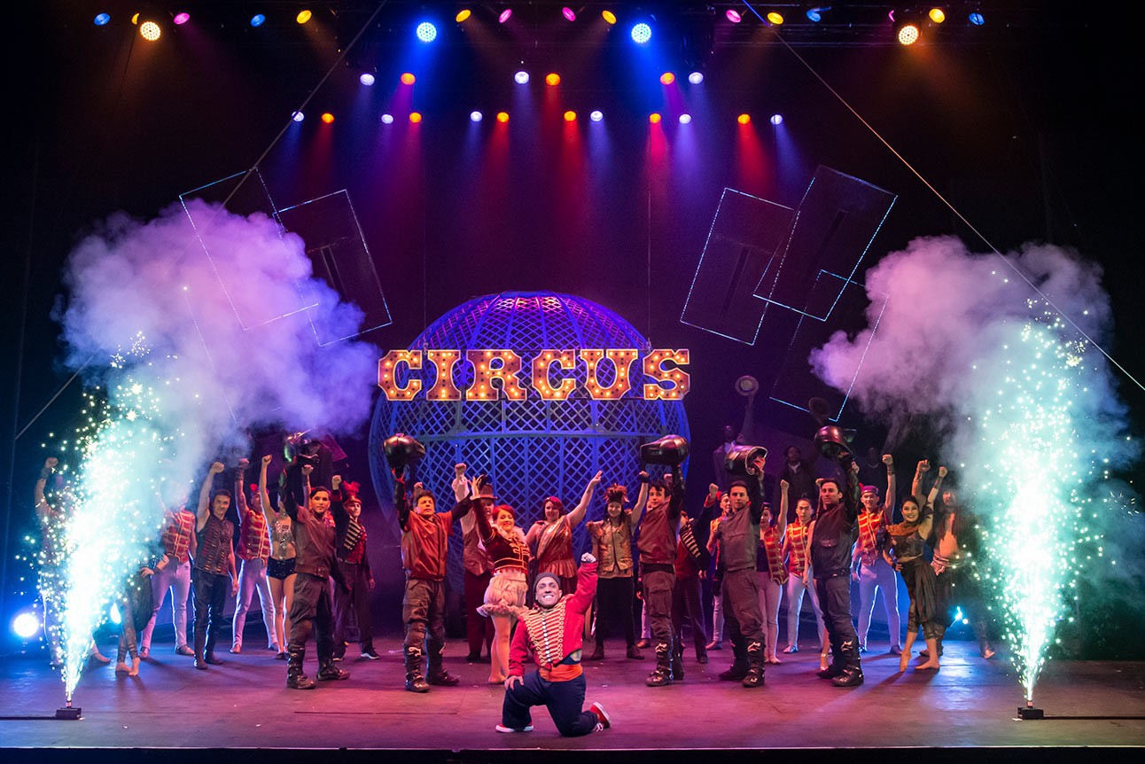 Performers in vibrant costumes on stage, colourful lights and smoke, with "CIRCUS" sign in background.