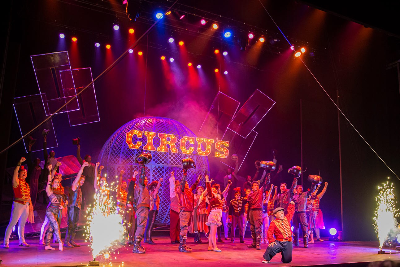 Performers in colourful costumes on stage, with "CIRCUS" sign lit up, vibrant lights and pyrotechnics.