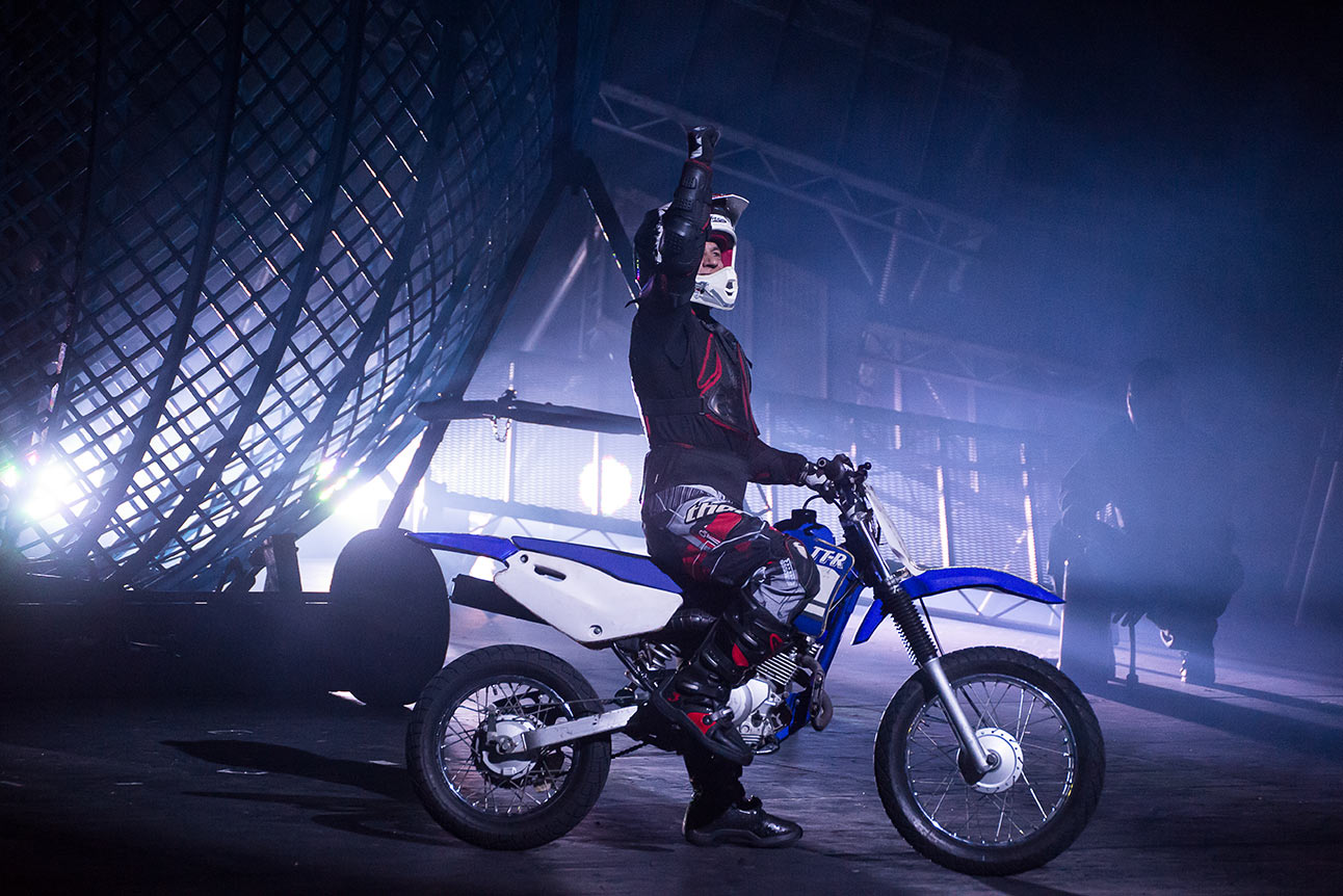 Motorcyclist in protective gear raises arm inside metal globe structure, dramatic lighting, blue hues.