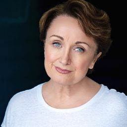 A woman with short brown hair wearing a white top, smiling softly against a dark background.
