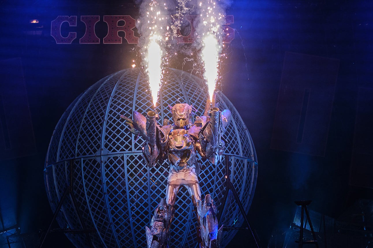 Performer in metallic costume emits sparks, standing before a spherical cage, with dramatic blue lighting.