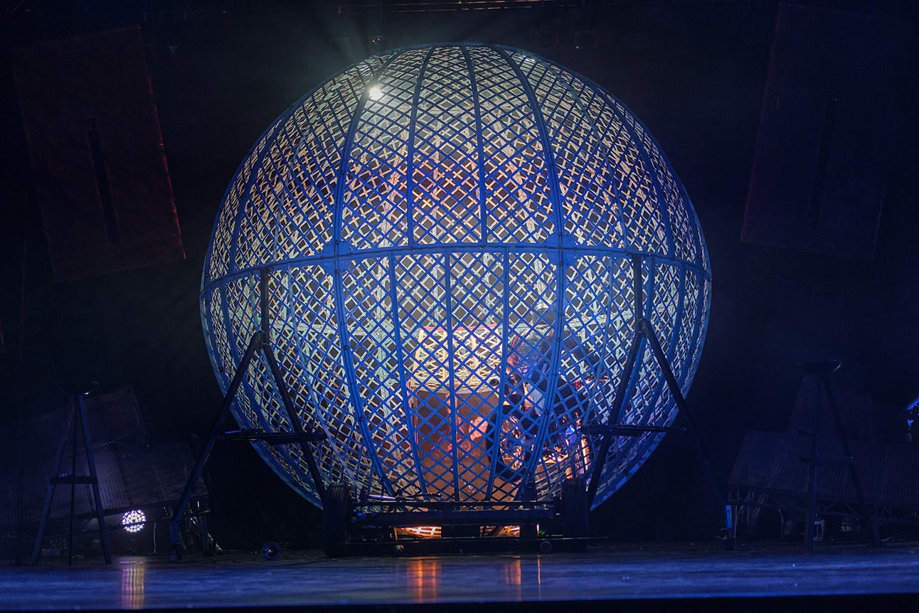 Motorcyclists performing inside a large metal globe at Cirque Berserk, illuminated with blue lighting.