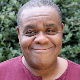 A man in a maroon shirt smiles warmly, with green foliage in the background.