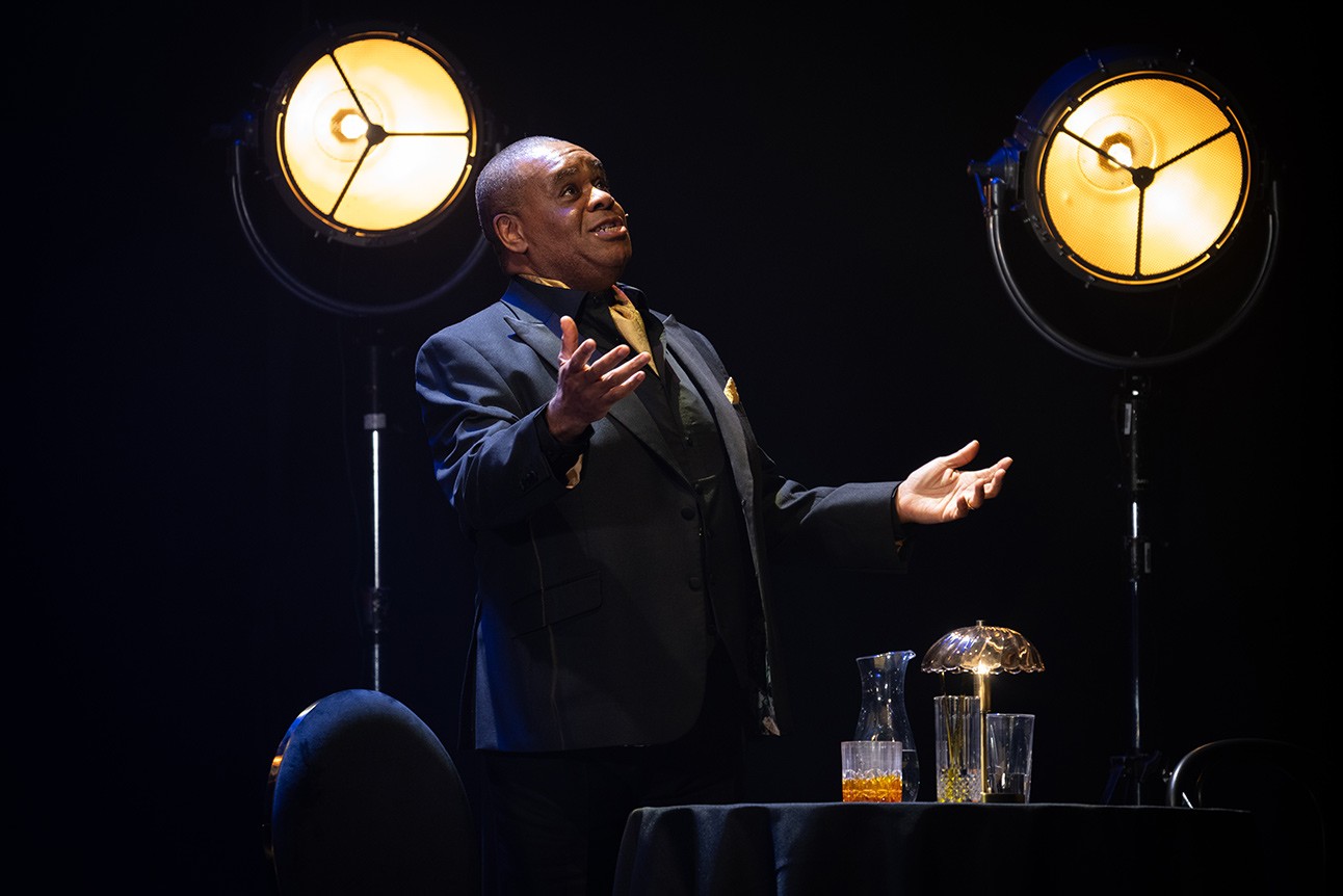 A man in a suit gestures expressively under stage lights, with a table and drinks nearby.