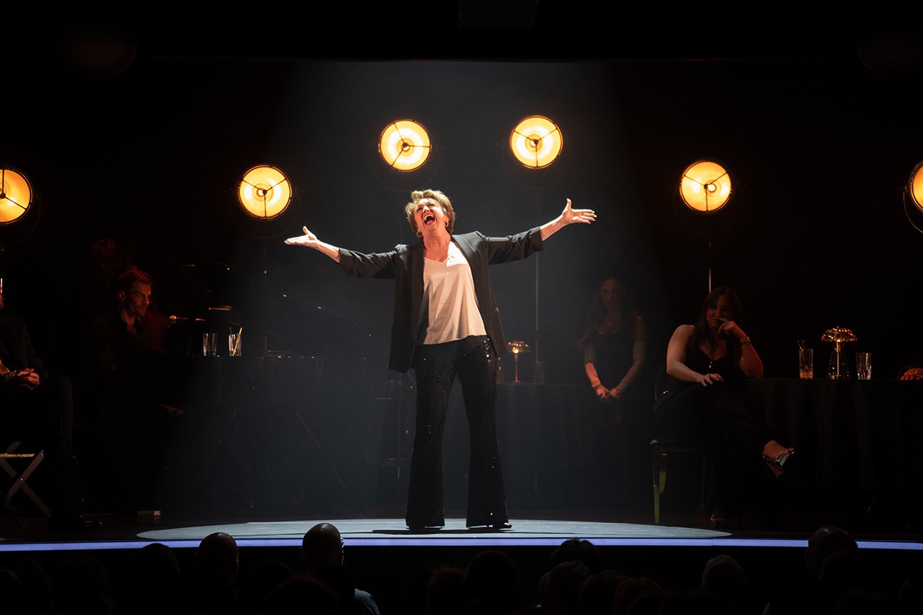 Performer on stage with arms outstretched, illuminated by four circular lights, dark background.