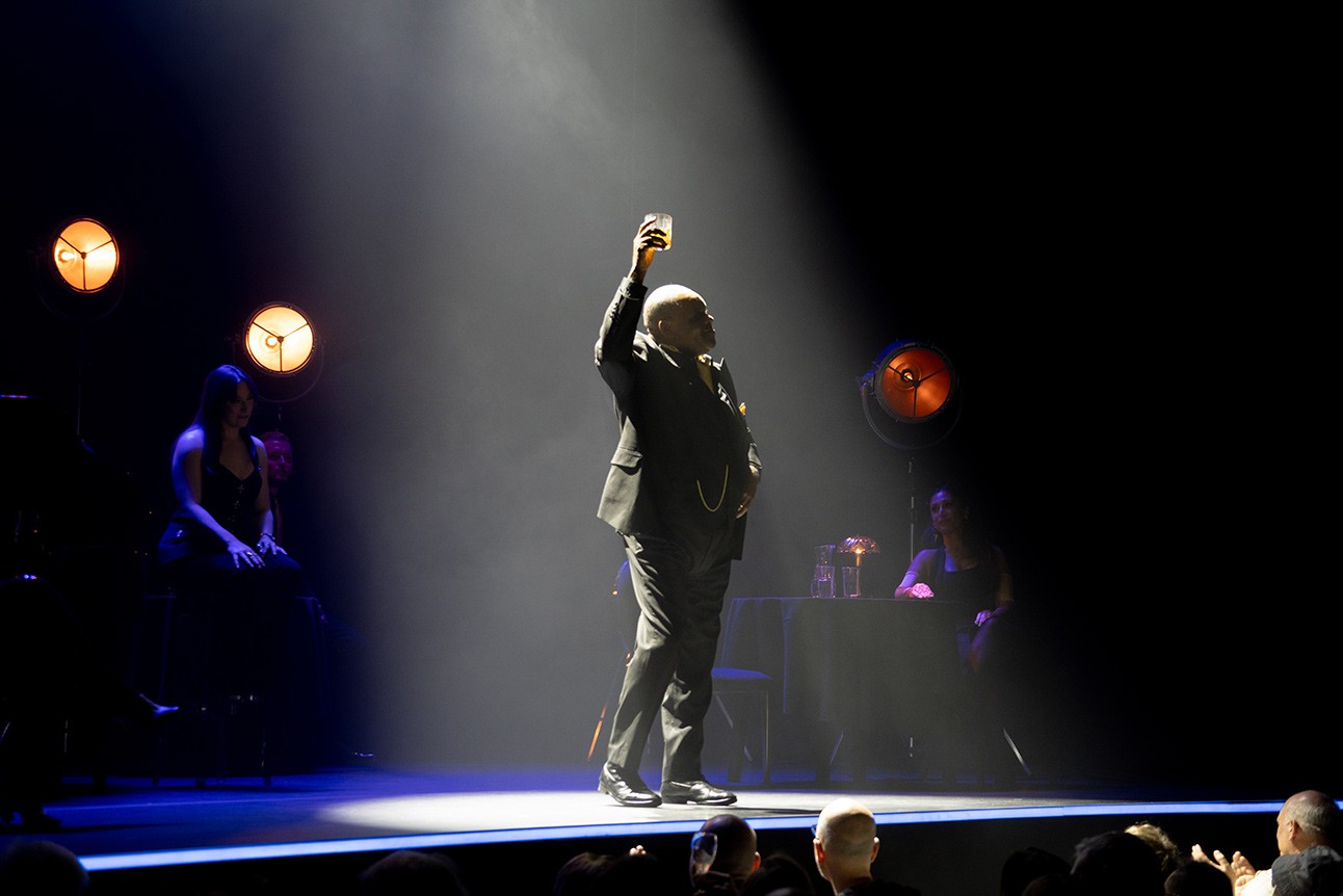 Performer in spotlight raises arm on dimly lit stage; audience and warm lights in background.
