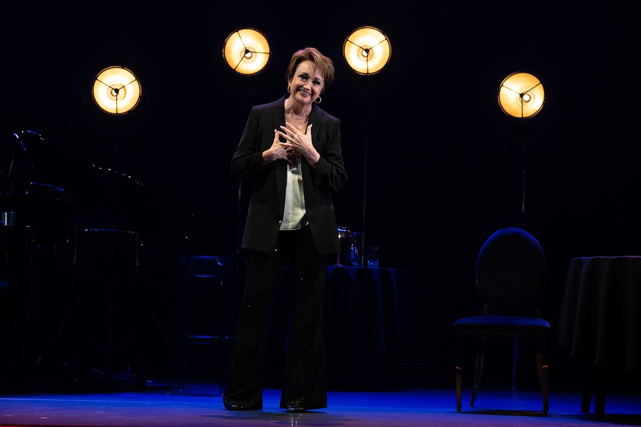 A person in a dark suit stands on stage, smiling, with four round lights behind them.