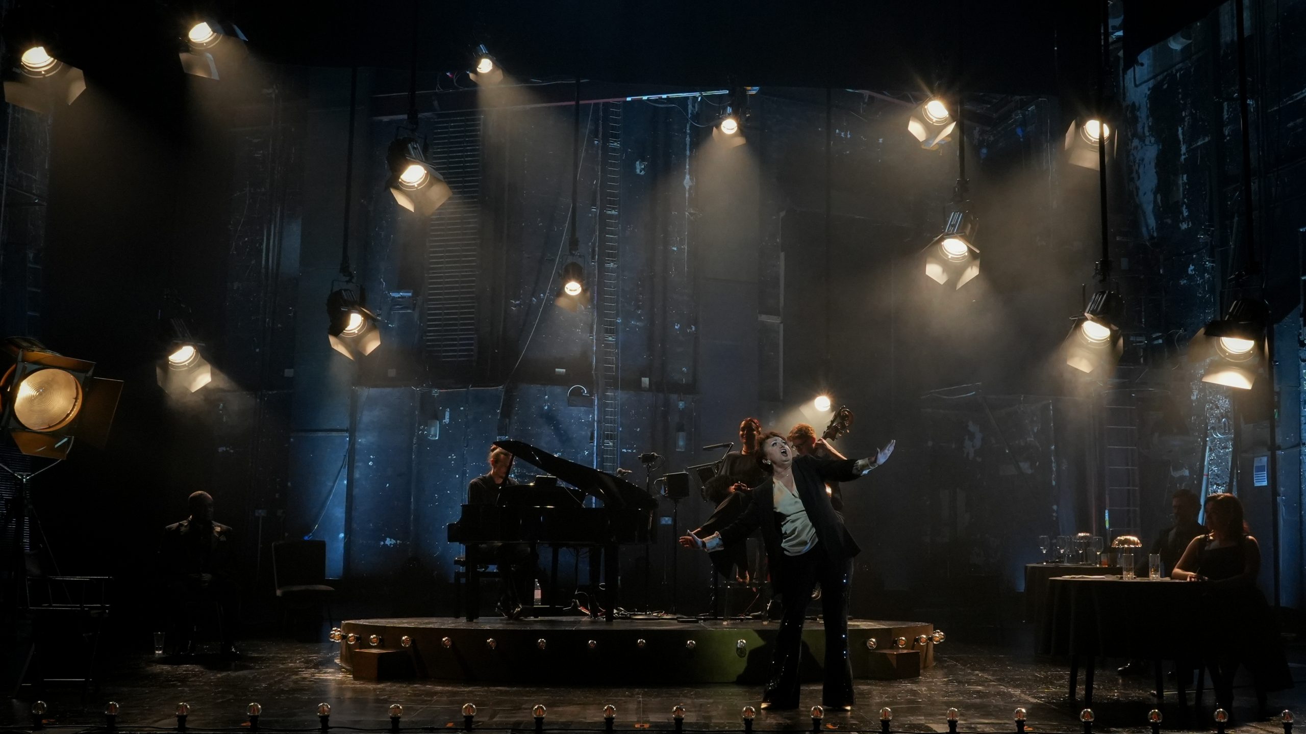 Performer in spotlight sings passionately on stage, pianist plays grand piano; dimly lit theatre backdrop.