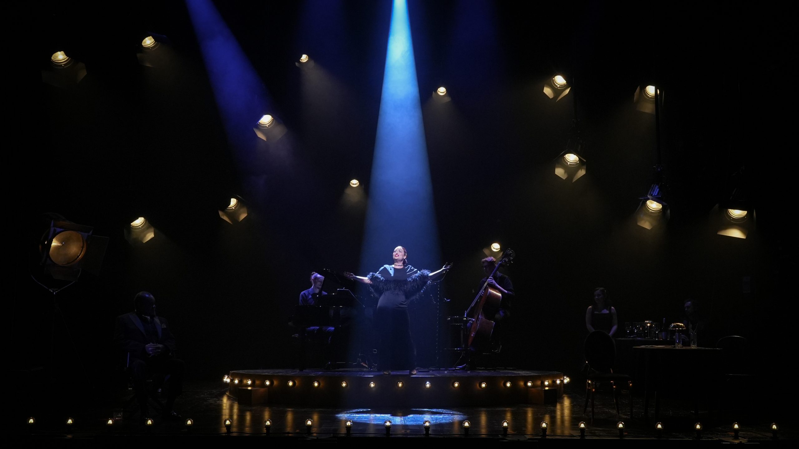 Performer stands centre stage under blue spotlight, arms outstretched, surrounded by musicians, dark background.