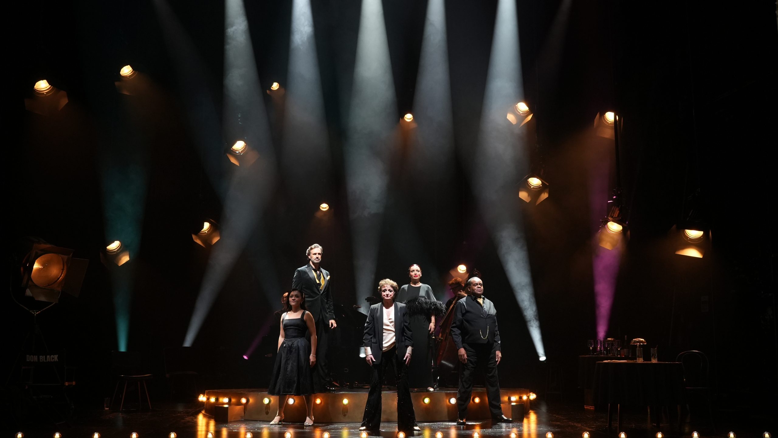 Performers in formal attire stand under spotlights on stage at the Garrick Theatre.