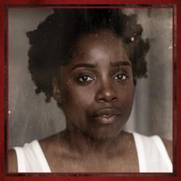 A woman with short curly hair wearing a white top, framed in a red border.
