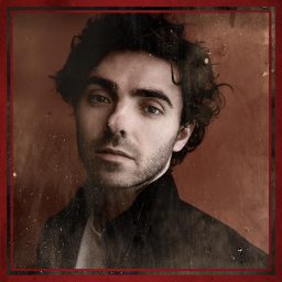 A man with dark hair and a serious expression, wearing a dark jacket, sepia-toned background.