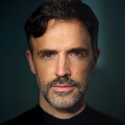 A man with short dark hair and a beard looks directly at the camera, dark background.