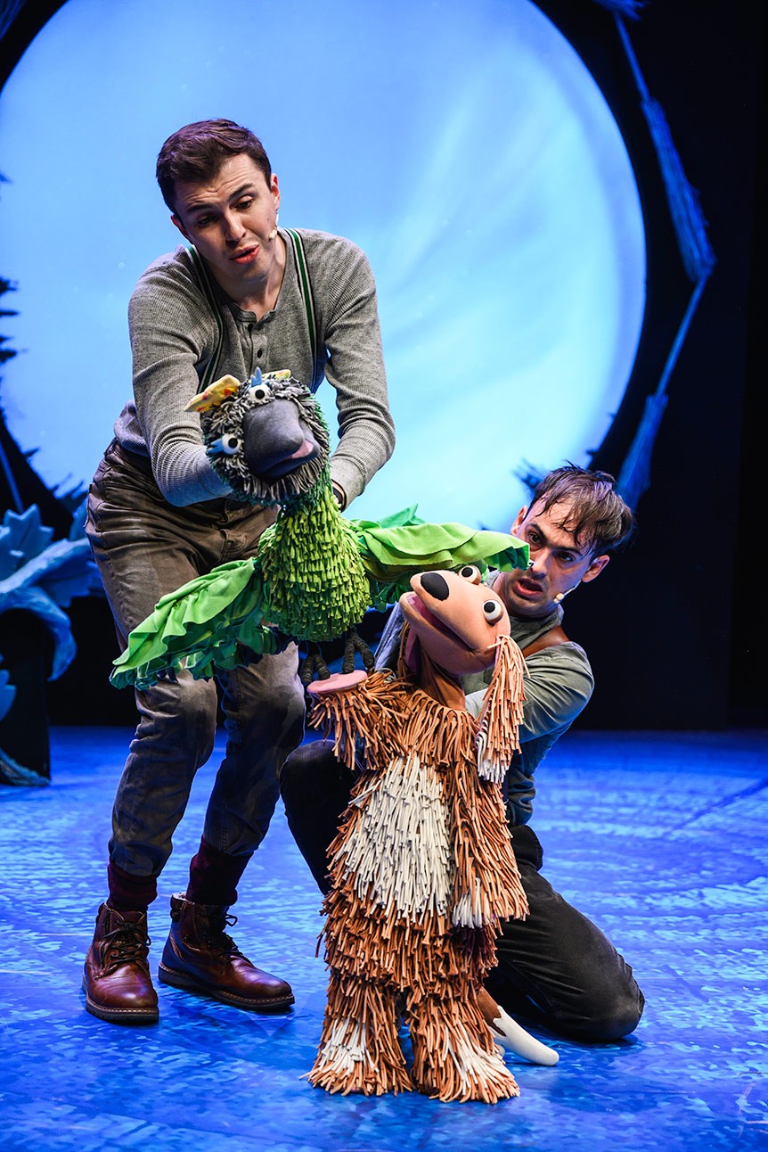 Two performers manipulate bird and dog puppets on stage, with a blue-lit circular backdrop.