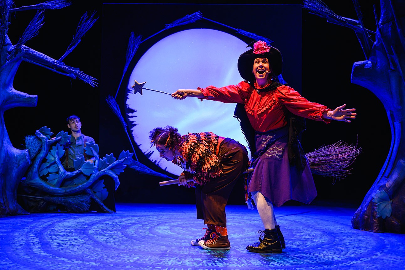 Performers on stage in colourful costumes, witch holding wand, large moon backdrop, theatrical setting.