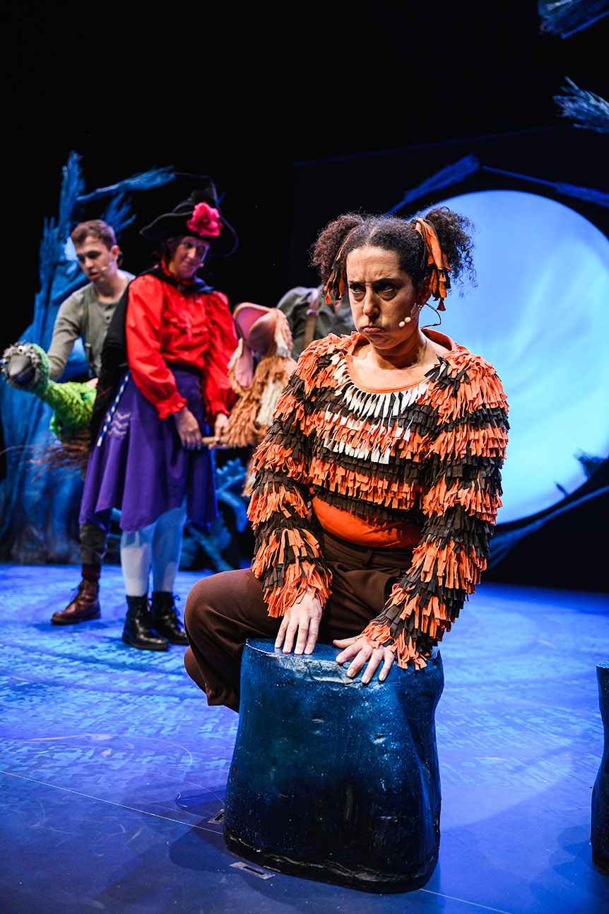 Performer in cat costume sits on stage, with actors in colourful outfits in the background.