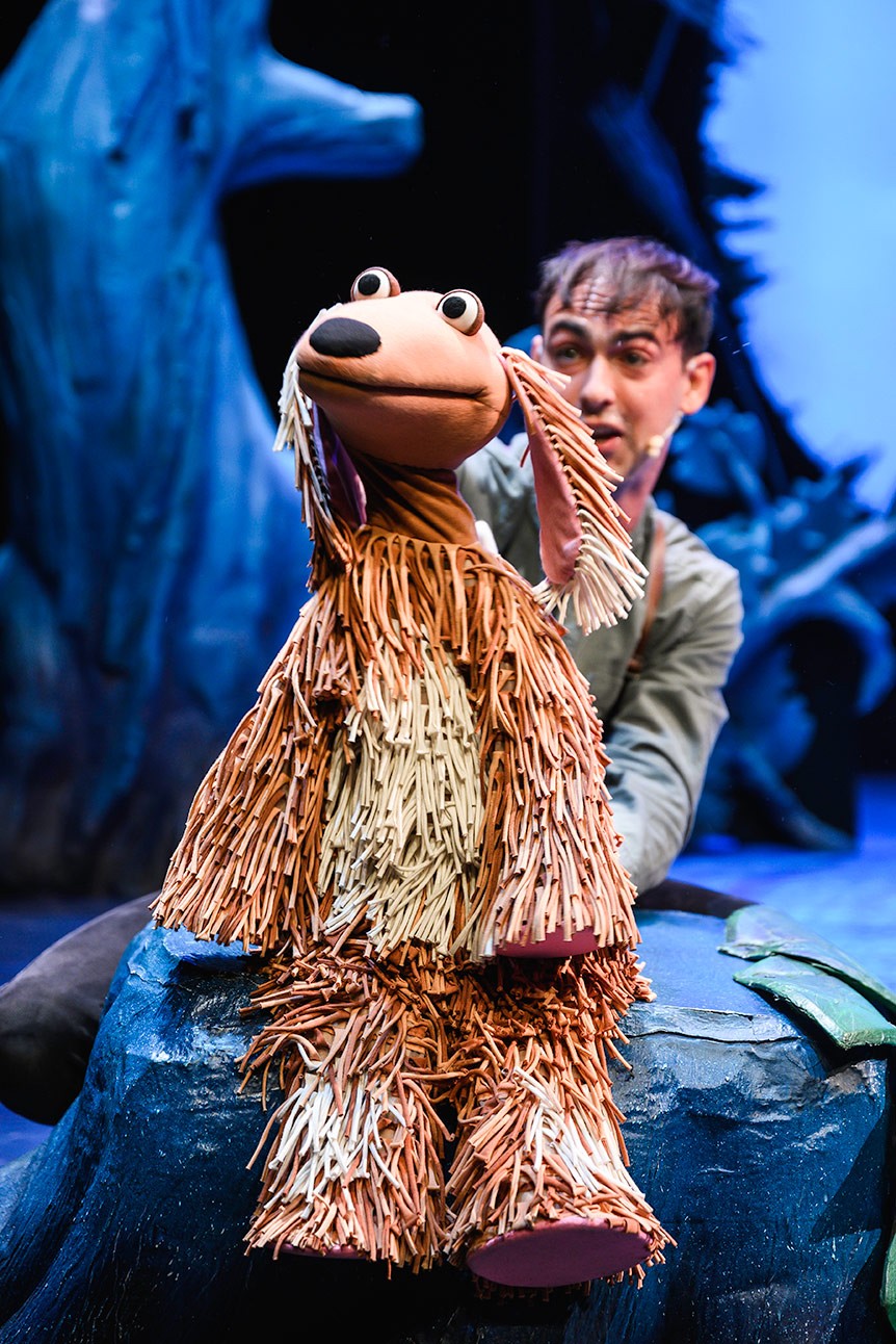 A performer manipulates a shaggy dog puppet on stage, with a blue and green backdrop.