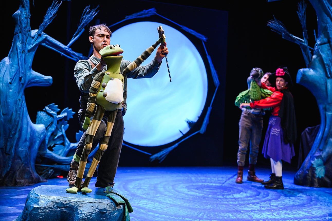 Performer manipulates frog puppet on stage; large moon backdrop, two others hold puppets in background.