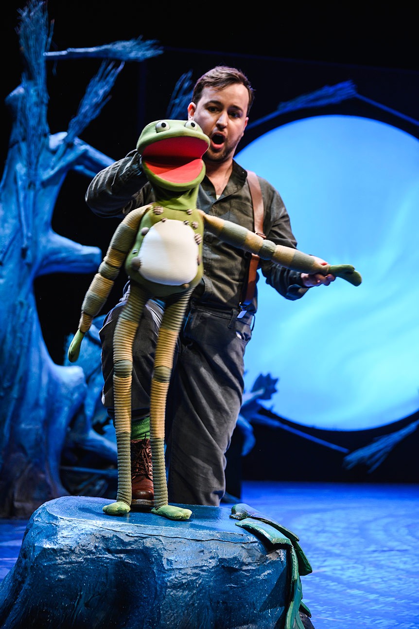 Performer operates a frog puppet on stage, with a blue-lit tree and circular backdrop behind.