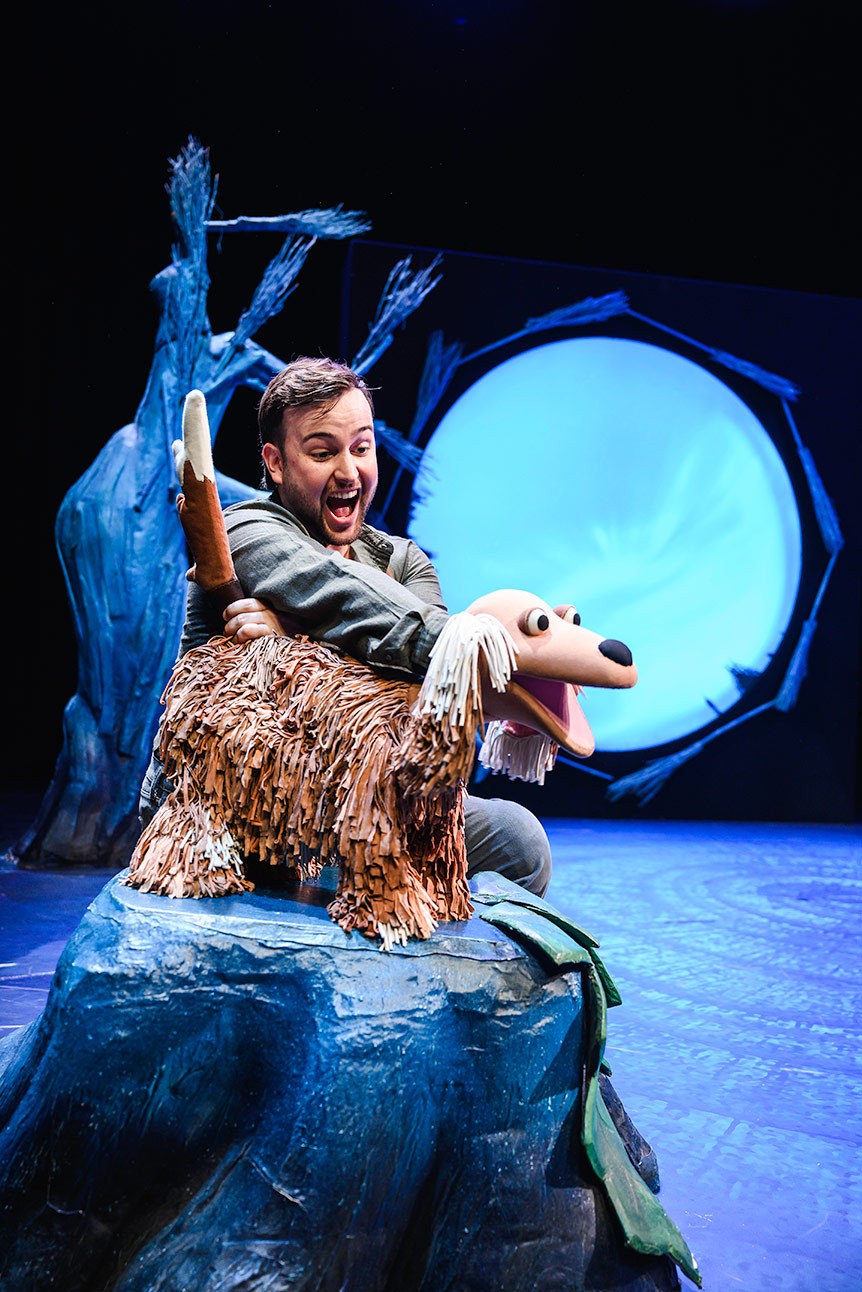 Actor enthusiastically interacts with a large dog puppet on stage, with a blue-lit backdrop.