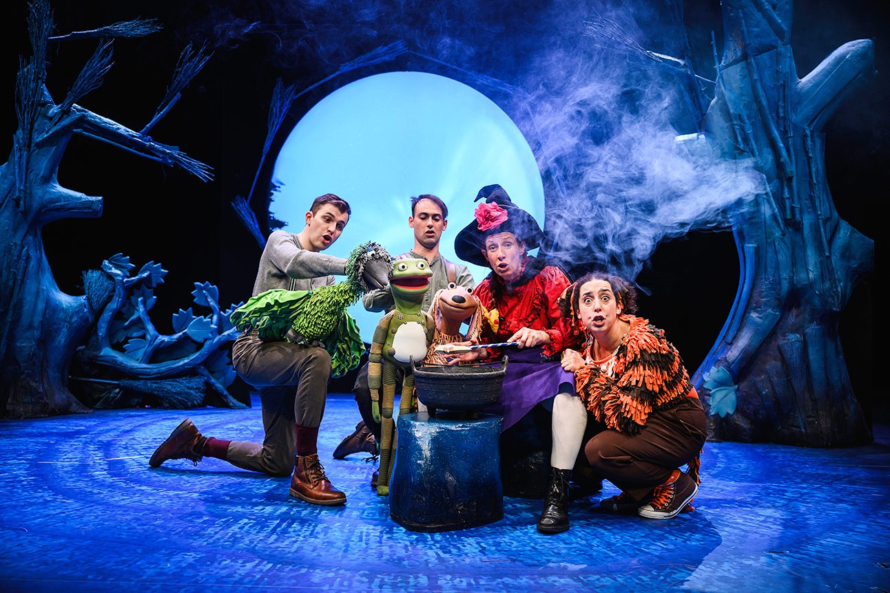 Five performers in colourful costumes gather around a steaming cauldron on a blue-lit stage.