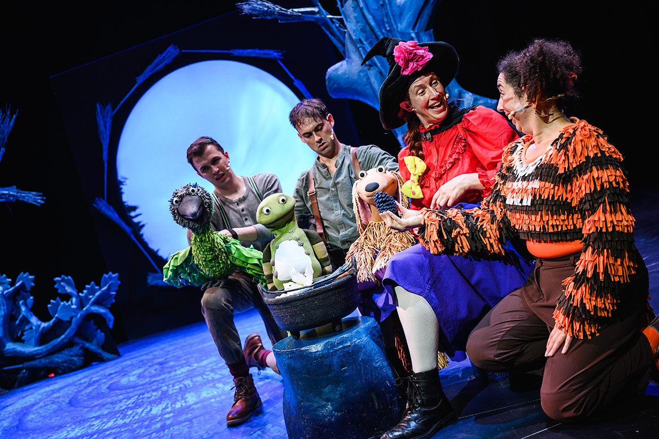 Performers with puppets around a cauldron on stage, vibrant costumes, blue-lit background.
