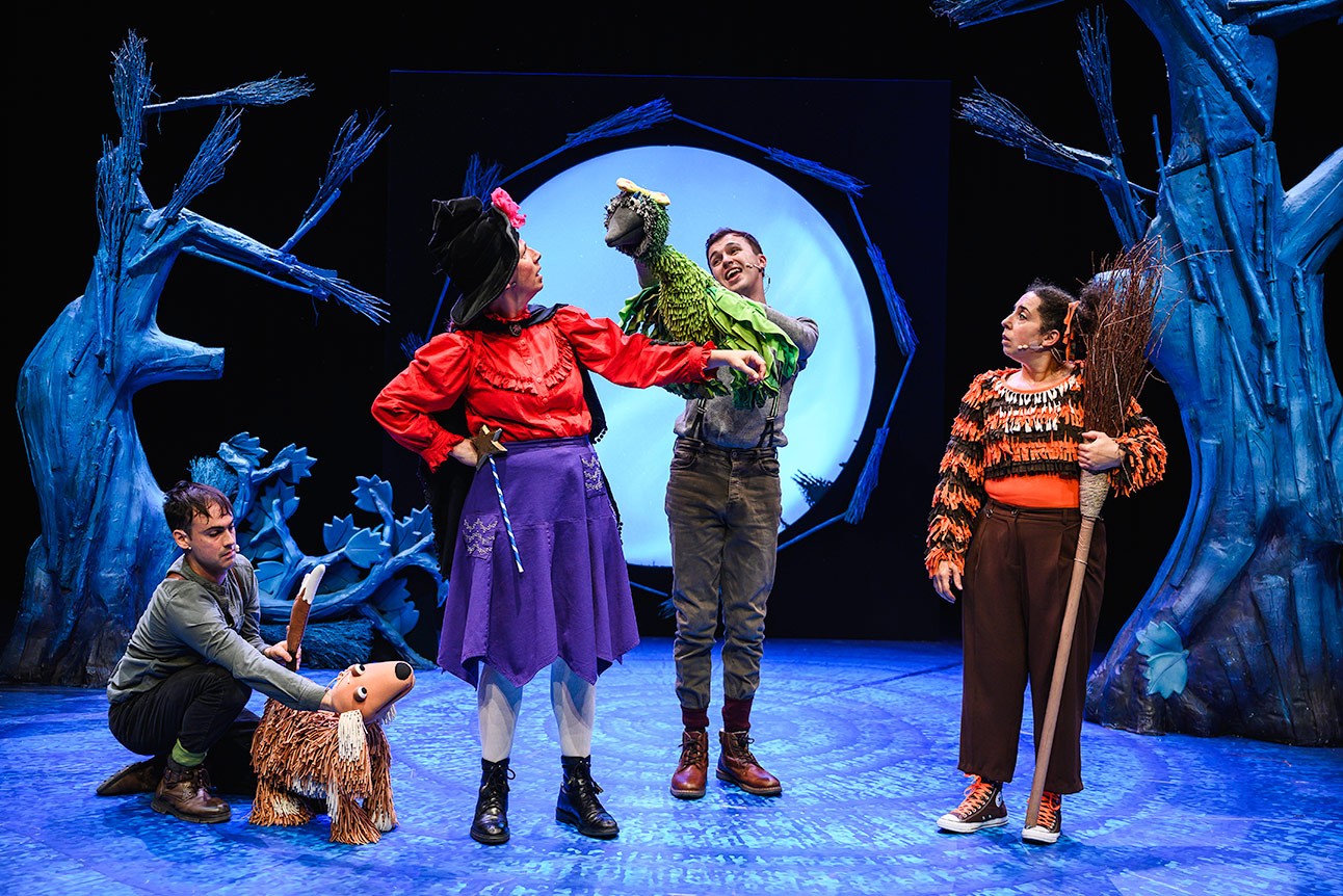 Performers on stage, one dressed as a witch with a green bird puppet on her arm.
