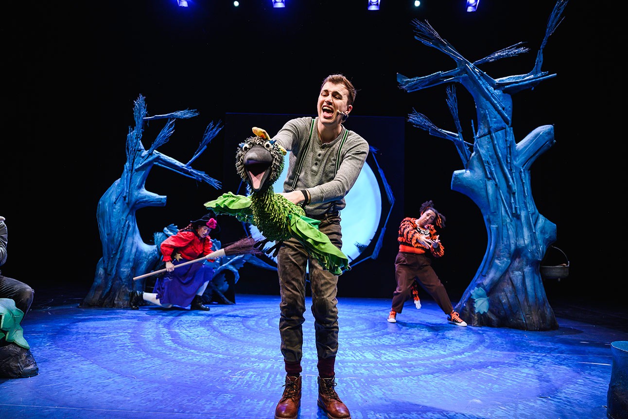 Performer holding green puppet bird on stage, with two actors in colourful costumes in background.