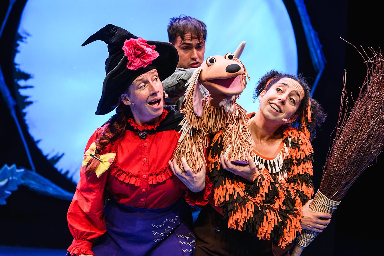 Performers in colourful costumes hold a dog puppet on stage, with a blue background.