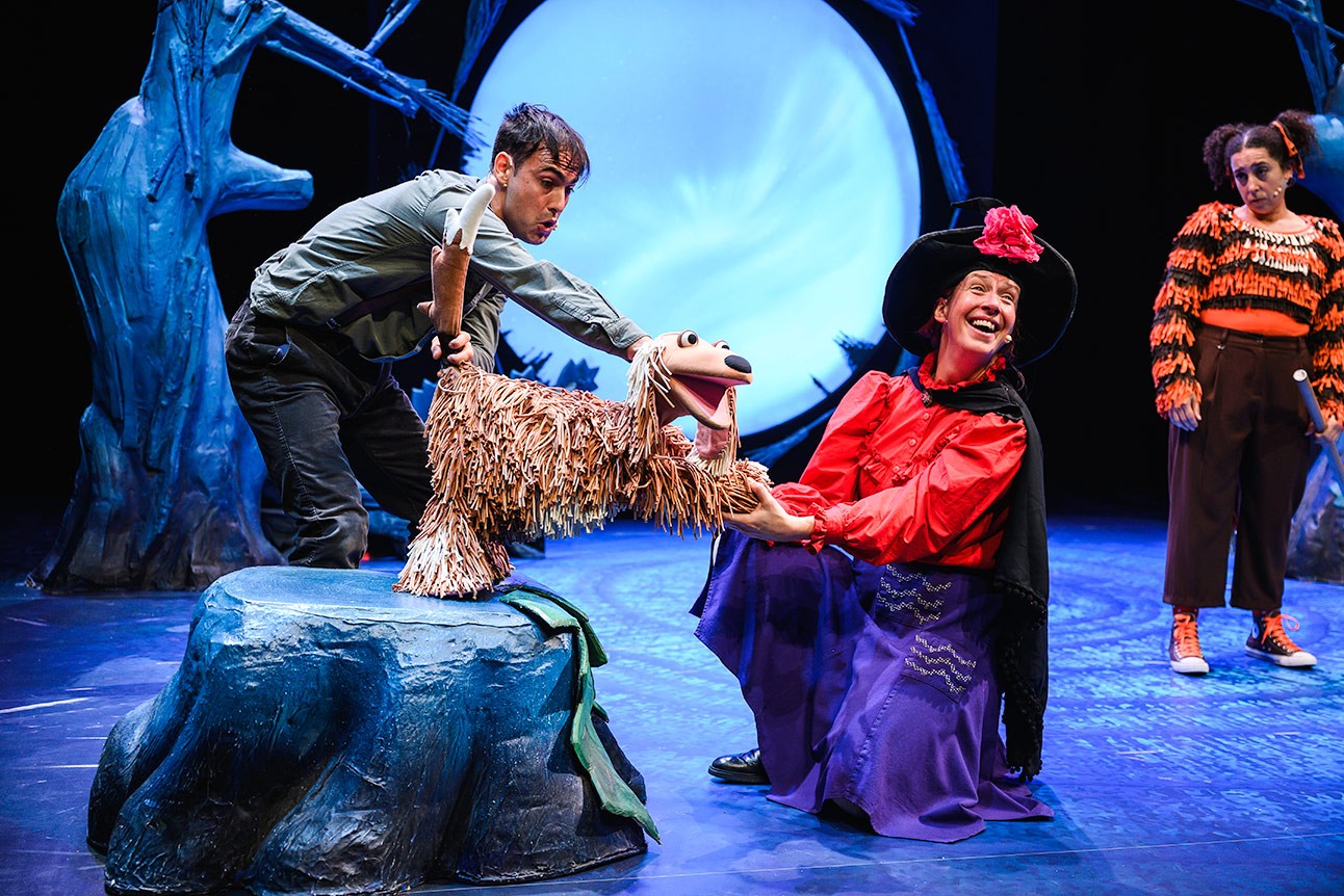 Performer in witch costume holds puppet dog's paw on stage, with blue-lit background.