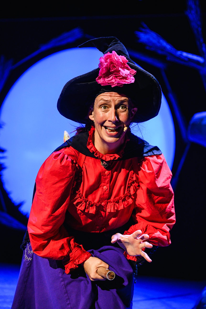 Performer dressed as a witch in red costume and black hat, smiling on stage with blue lighting.