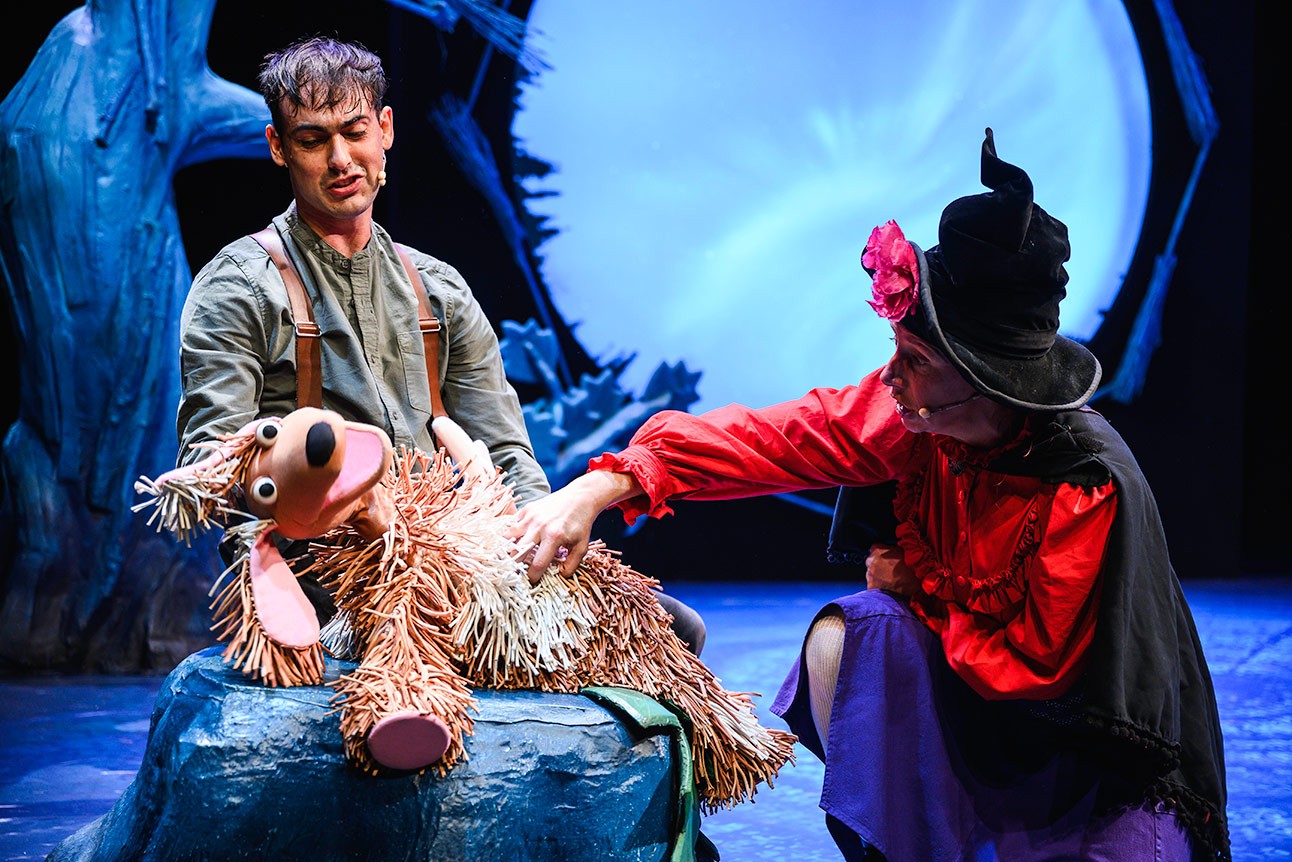 A witch in a black hat tickles a large, smiling dog puppet on stage.